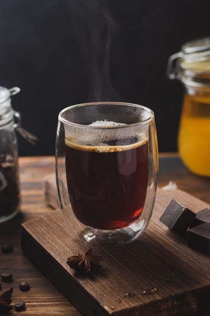 Café noir dans le verre avec de la vapeur sur une surface en bois, du miel et du chocolat noir