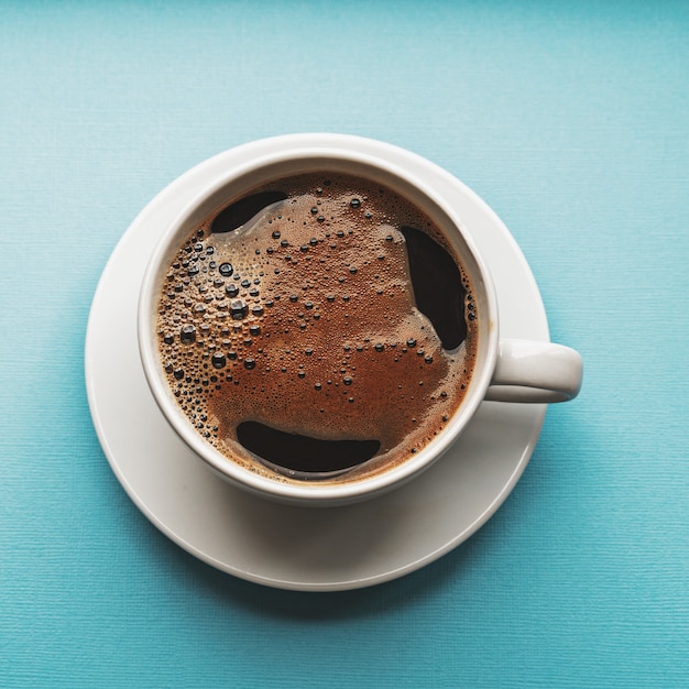 Café noir avec mousse dans une tasse blanche sur fond bleu, mise à plat.