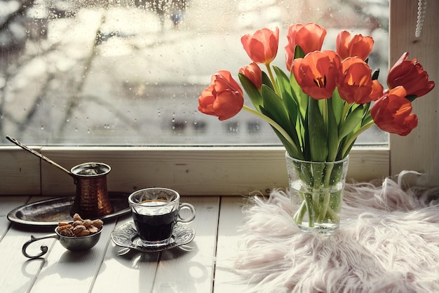 Café oriental dans une cafetière turque traditionnelle en cuivre avec des fleurs sur le rebord de la fenêtre. Rebord de fenêtre en bois rustique avec bouquet de tulipes et livre. Jour de pluie froide au printemps. Scène confortable, concept hygge.