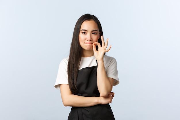 Café, petite entreprise et concept de démarrage. Barista coréen mignon et sérieux, employée au café promet de garder le secret, de fermer les lèvres ou de verrouiller les lèvres, debout dans un tablier noir