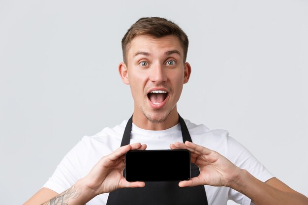 Café et restaurants propriétaires de café et concept de vente au détail excité et étonné beau vendeur en tablier noir bouche ouverte amusé et montrant l'application smartphone affichage mur blanc