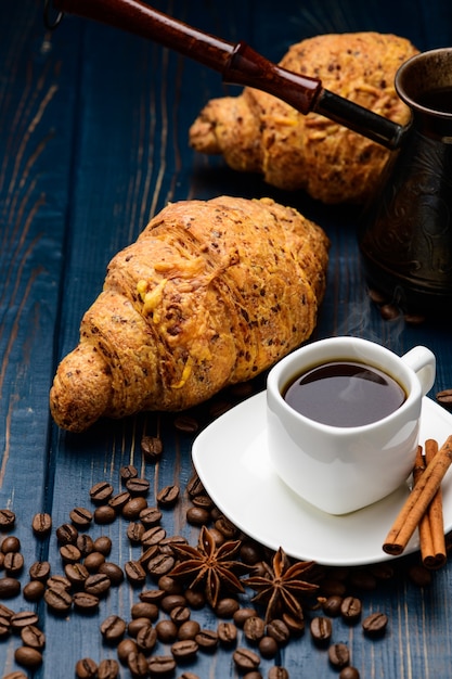 Le café se déverse dans une tasse sur une table en bois bleue avec des grains de café et un croissant.