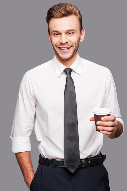 Café et travail. Gai jeune homme tenant une tasse de café en se tenant debout sur fond gris