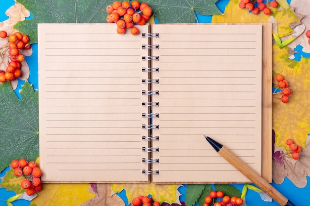 Cahier d'artisanat et stylo ligné vierge à plat sur les feuilles colorées sèches tombées à l'automne sur fond bleu. Étudier à l'école en automne en septembre et octobre concept. Modèle de maquette de bloc-notes vierge