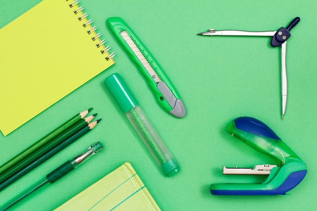 Cahier, crayons de couleur, stylo, livre, feutre, coupe-papier, boussole et agrafeuse sur fond vert. Vue de dessus. Retour au concept de l'école. Fournitures scolaires