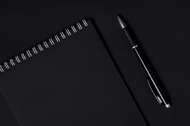 Cahier avec feuilles noires et reliure à spirale blanche et stylo noir sur fond sombre, vue de dessus, espace de copie. Concept minimal d'entreprise ou d'éducation