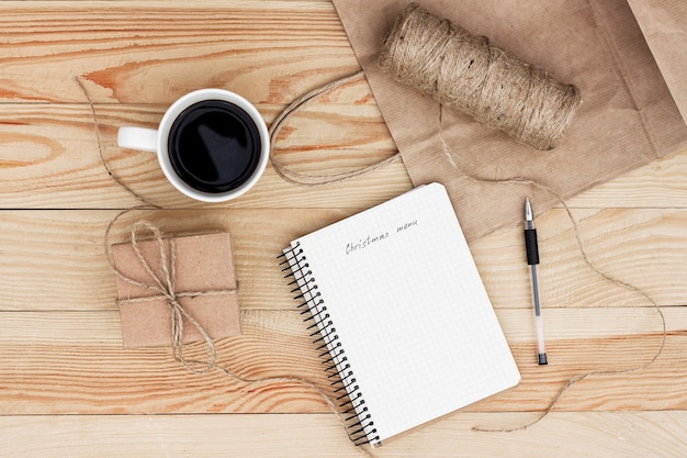 Un cahier avec une inscription Liste de menus de Noël posée sur une table en bois. Il est entouré d'un stylo noir, d'une tasse de café, d'un cadeau, d'une ficelle et d'un sac en papier.