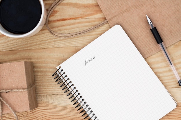 Un cahier avec une inscription Menu portant sur une table en bois. Il est entouré d'un stylo noir, d'une tasse de café, d'un cadeau et d'un sac en papier.