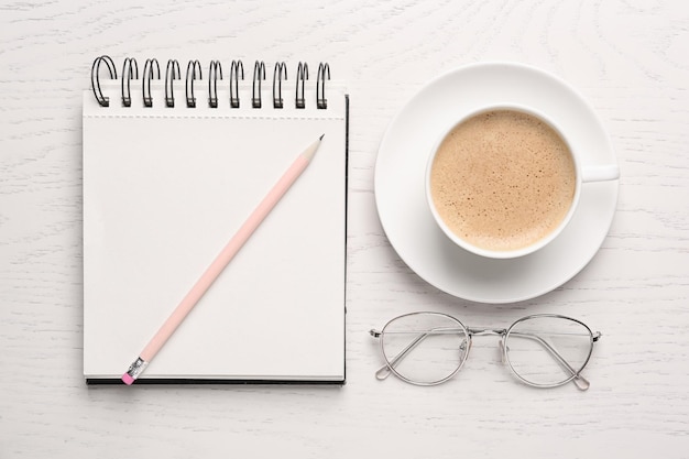 Cahier avec des lunettes à crayons et une tasse de café sur une table en bois blanche à plat