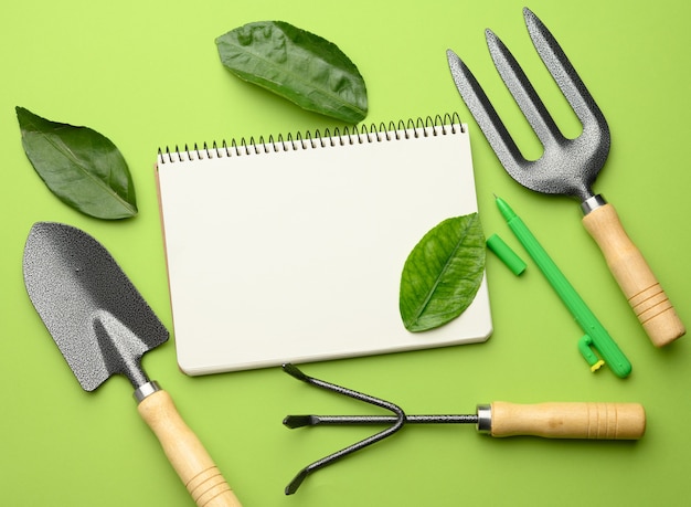 Cahier ouvert avec des feuilles blanches vierges et divers outils de jardinage avec des poignées en bois sur un mur vert, à plat, copiez l'espace