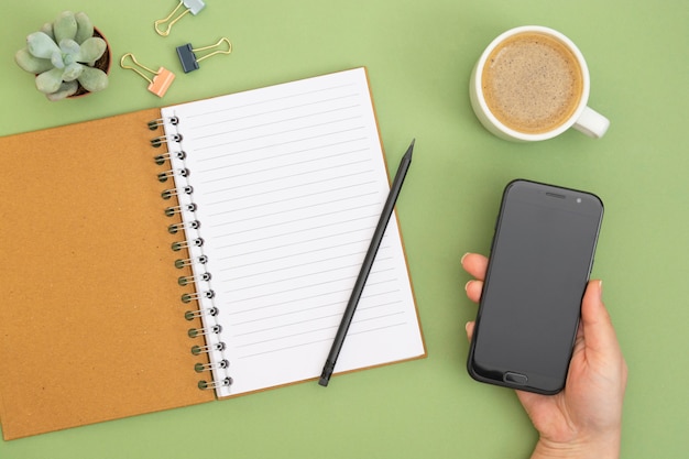 Cahier ouvert avec page vide, crayon, tasse à café et main tenant un smartphone. Plateau de table, espace de travail sur fond vert. Mise à plat créative.