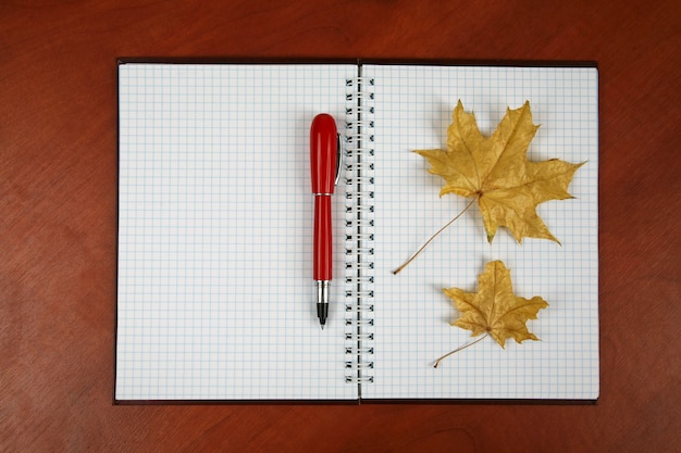 Le cahier ouvert et le stylo rouge avec la feuille d'automne se trouvant sur une table en bois
