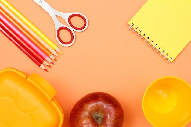 Cahier, pomme, ciseaux, crayons de couleur, gobelet en plastique et boîte à lunch sur fond rose.