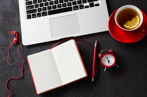 Cahier rouge vierge, ordinateur portable, réveil, casque et tasse de thé