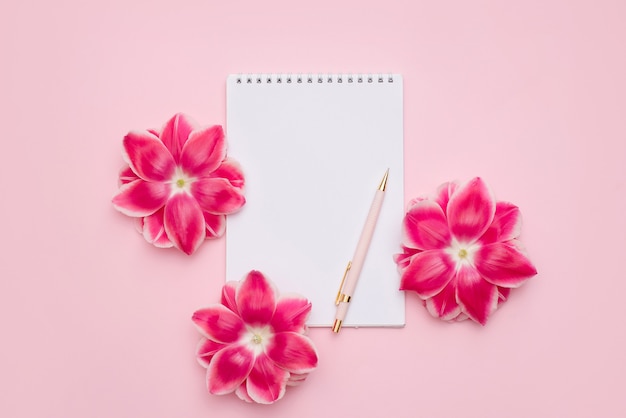 Cahier sur une spirale avec une feuille blanche vierge, un stylo et des fleurs roses sur une surface rose clair