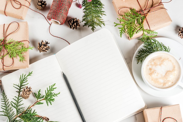 Cahier vierge et crayon pour souhaits, liste de choses à faire, tasse à café, cadeau de Noël ou boîte cadeau, décoré de branches d'arbres de Noël, pommes de pin, fruits rouges, sur une table en marbre blanc, copie vue de dessus de l'espace