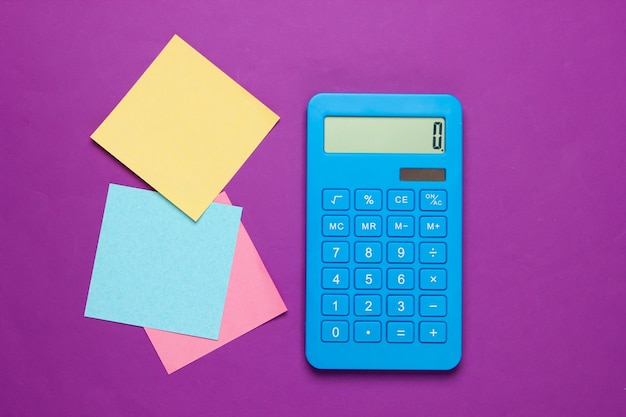 Calculatrice bleue avec des feuilles de papier mémo colorées sur violet. Outils bureautiques