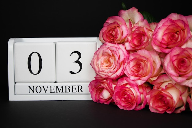 Calendrier en bois du 3 novembre, blanc sur fond noir, des roses roses se trouvent à proximité.