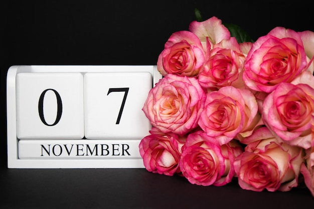 Calendrier en bois du 7 novembre, blanc sur fond noir, des roses roses se trouvent à proximité.