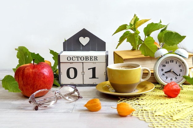 Calendrier du 1er octobre une maison décorative avec les chiffres 01 le nom du mois octobre en anglais une pomme rouge des verres des branches de physalis une pile de livres une tasse de café une horloge