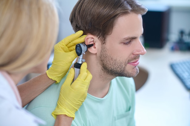 Calme beau jeune homme de race blanche barbu aux cheveux noirs examiné par une oto-rhino-laryngologiste professionnelle en gants stériles