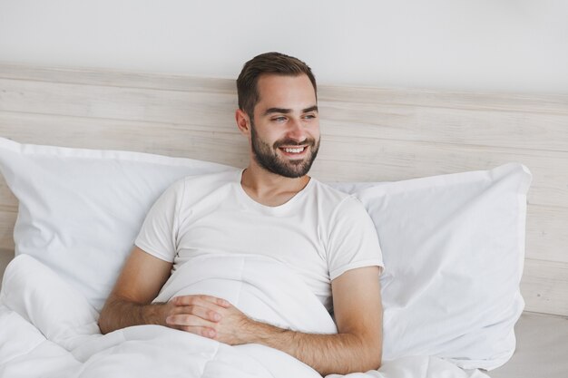 Calme jeune bel homme barbu allongé dans son lit avec une couverture d'oreiller en drap blanc dans la chambre à la maison