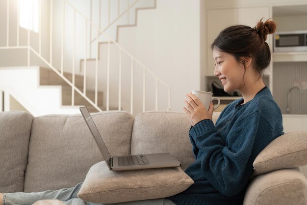 Calme jeune femme asiatique travaillant sur un ordinateur portable assis sur un canapé confortable fille heureuse naviguant sur Internet ou faisant des achats en ligne pendant le week-end ensoleillé à la maison se détendre sur un canapé avec ordinateur