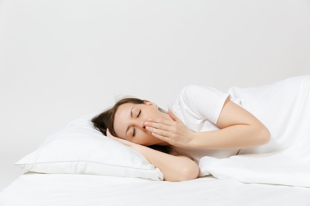 Calme jeune femme brune allongée dans son lit avec drap blanc, oreiller, couverture sur mur blanc