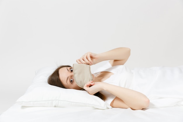 Calme jeune femme brune allongée dans son lit avec masque de sommeil sur drap blanc, oreiller, couverture sur mur blanc