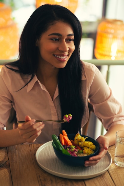 Calme jolie dame assise dans un café seule et souriante tout en tenant une fourchette et en mangeant de la salade