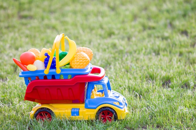 Camion de voiture de jouet coloré en plastique brillant transportant un panier avec fruits et légumes jouet à l'extérieur sur une journée d'été ensoleillée.