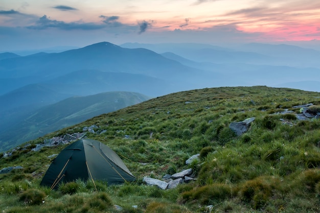 Camping d'été dans les montagnes à l'aube. Tente touristique sur une colline herbeuse ronde sur des montagnes bleues brumeuses éloignées sous le ciel rose avant le lever ou le coucher du soleil. Tourisme, randonnée et beauté du concept de la nature.