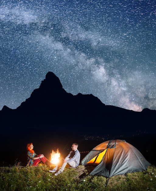 Camping de nuit. Couple heureux backpackers assis devant un feu de joie et tente sous un ciel étoilé incroyablement beau. Silhouette des hautes montagnes et village dans la vallée en arrière-plan. Longue exposition