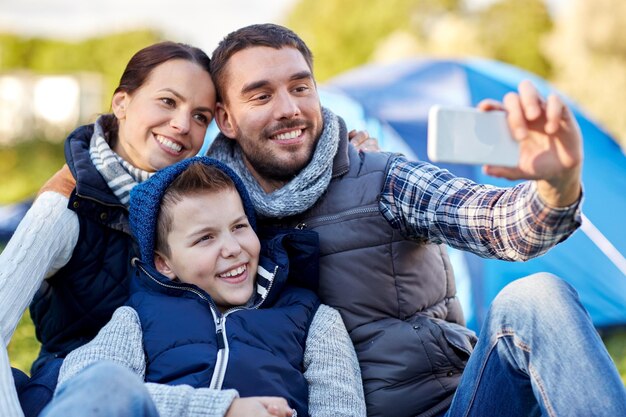 camping, randonnée, technologie et concept de personnes - famille heureuse avec smartphone prenant selfie au camping