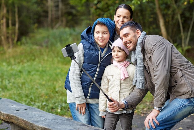 camping, tourisme, randonnée, technologie et concept humain - famille heureuse prenant des photos avec un smartphone sur un bâton de selfie au camp