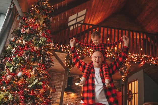 Candide authentique père heureux et son fils en pyjama à carreaux rouges s'amusant au pavillon en bois décoré de Noël