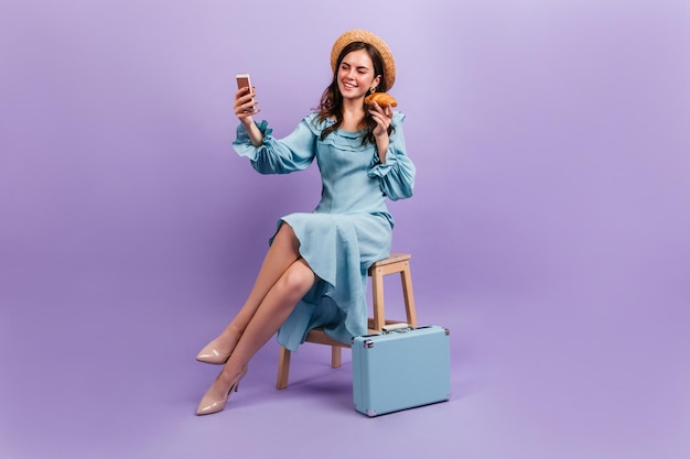 Un canotier parisien fait de beaux sourires et fait du selfie Une fille élégante tenant un croissant appétissant et assise à côté d'une valise rétro