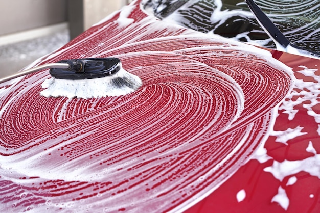 Capot avant de voiture rouge lavé dans un lave-auto en libre-service, détail sur la brosse laissant des traits dans de la mousse de savon blanche.