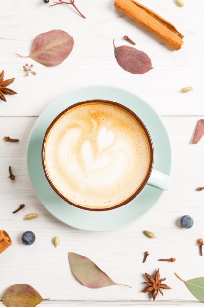 Cappuccino épicé d'automne. Tasse à café bleue avec mousse, feuilles sèches, clous de girofle, bâton de cannelle et étoiles d'anis au bois blanc. Concept d'offre saisonnière de boissons chaudes d'automne, de café et de bar. Vue de dessus