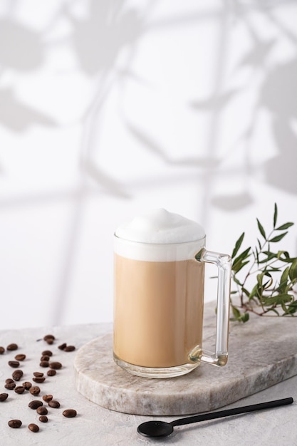 Cappuccino ou latte avec de la mousse de lait dans une tasse avec des grains de café et une cuillère sur fond de marbre clair avec ombre du matin