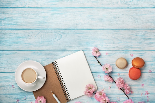 Cappuccino tasse blanche avec des fleurs de sakura, cahier, macarons, sur une table en bois bleue