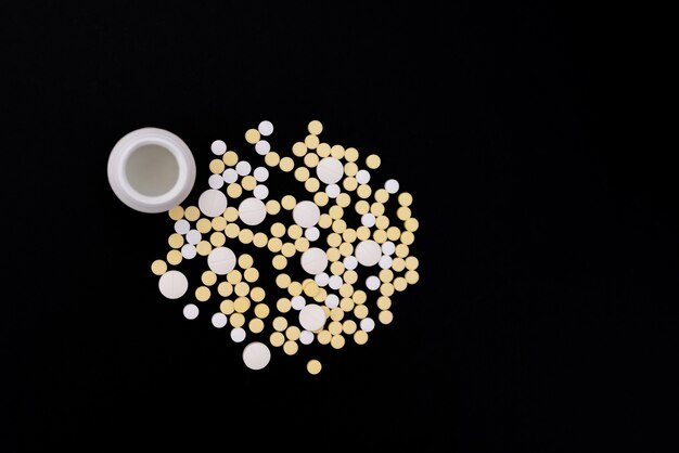 Capsules et comprimés de couleur blanche et jaune, un pot. Fond noir. Notion de santé