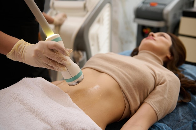 Capteur à ultrasons tactile en gros plan se déplaçant sur le ventre lubrifié au gel de contact d'une femme floue recevant une thérapie de cavitation de remodelage corporel dans une clinique de spa Soins corporels anticellulite professionnels