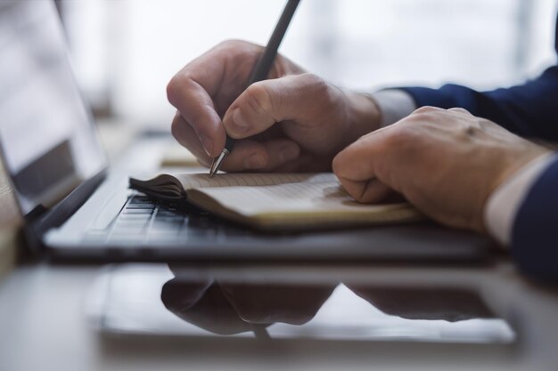 Capture en gros plan de la main d'un homme alors qu'il écrit dans un bloc-notes sur un ordinateur portable élégant avec un arrière-plan flou