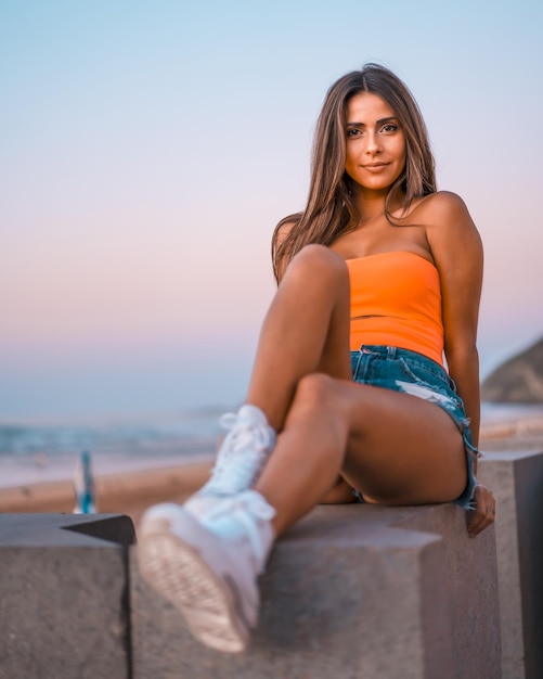 Capture verticale d'une jeune femme brune en short en jean et un haut orange assis sur la plage