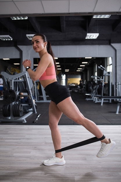 Capture verticale pleine longueur d'une belle sportive heureuse faisant des levées de jambes avec une bande de résistance à la salle de sport