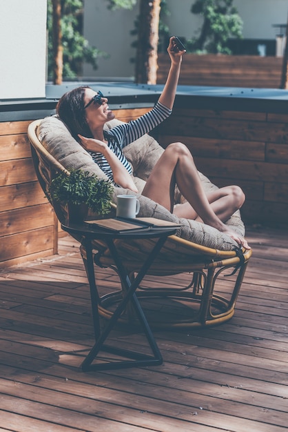 Capturer de grands moments. Belle jeune femme se reposant dans une grande chaise confortable sur sa terrasse extérieure de maison et faisant le selfie