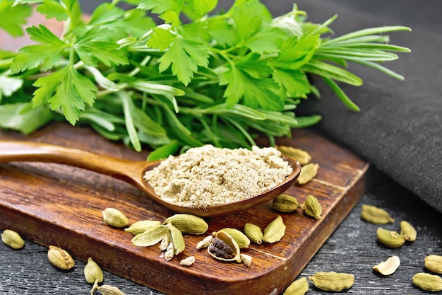 Cardamome Moulue Dans Une Cuillère, Capsules D'assaisonnement, Serviette, Persil Frais Et Romarin Sur Fond De Planche De Bois Noir