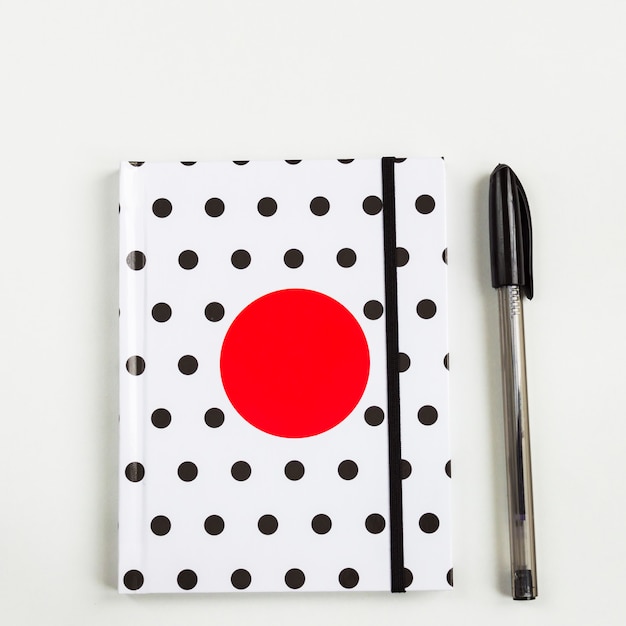 Carnet de notes à pois noir et blanc avec un cercle rouge sur la couverture et un stylo noir sur le tableau blanc. vue de dessus, pose à plat minimale