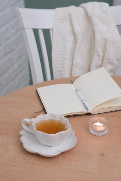 Un carnet ouvert avec un stylo blanc, une tasse de thé et une bougie allumée sur une table en bois et une chaise.